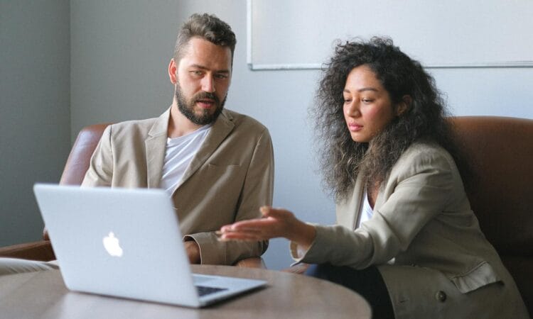 Serious colleagues in stylish outfits sitting at table and using laptop while discussing new project in office