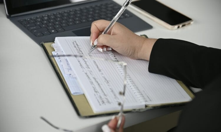 A person writing on a notebook with a laptop in the background
