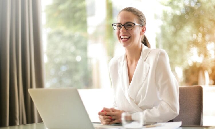 Cheerful businesswoman in glasses working on a laptop, in a bright and modern office setting.