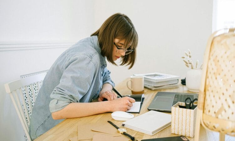 A young woman working from home, writing notes at a desk with a laptop and papers.