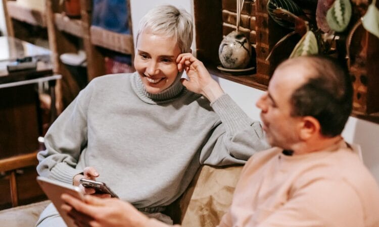 From above of middle aged diverse in casual clothes smiling while sitting on sofa and using smartphone and tablet during weekend at home