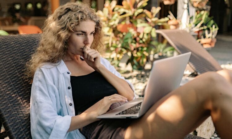 Woman with curly hair working remotely on a laptop while lounging outdoors in a tropical setting.
