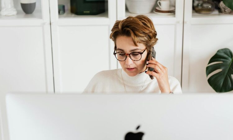 Smart woman with mobile in hand listening attentively to interlocutor and looking down while sitting in front of modern computer