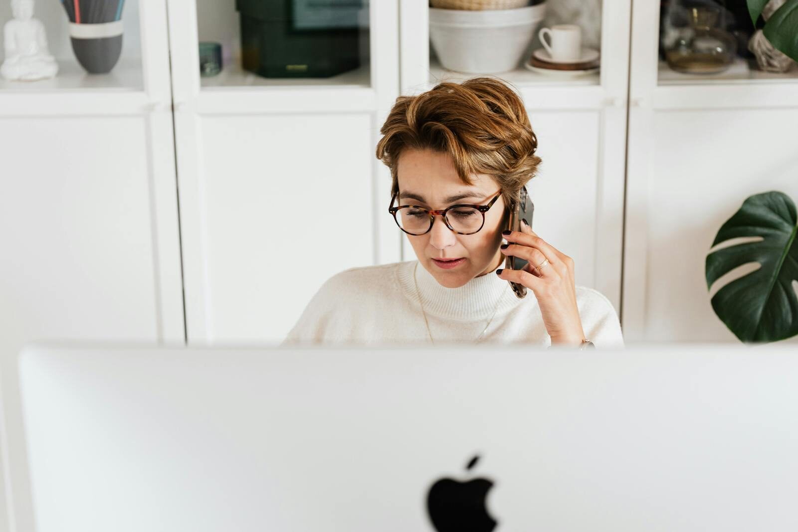 Smart woman with mobile in hand listening attentively to interlocutor and looking down while sitting in front of modern computer